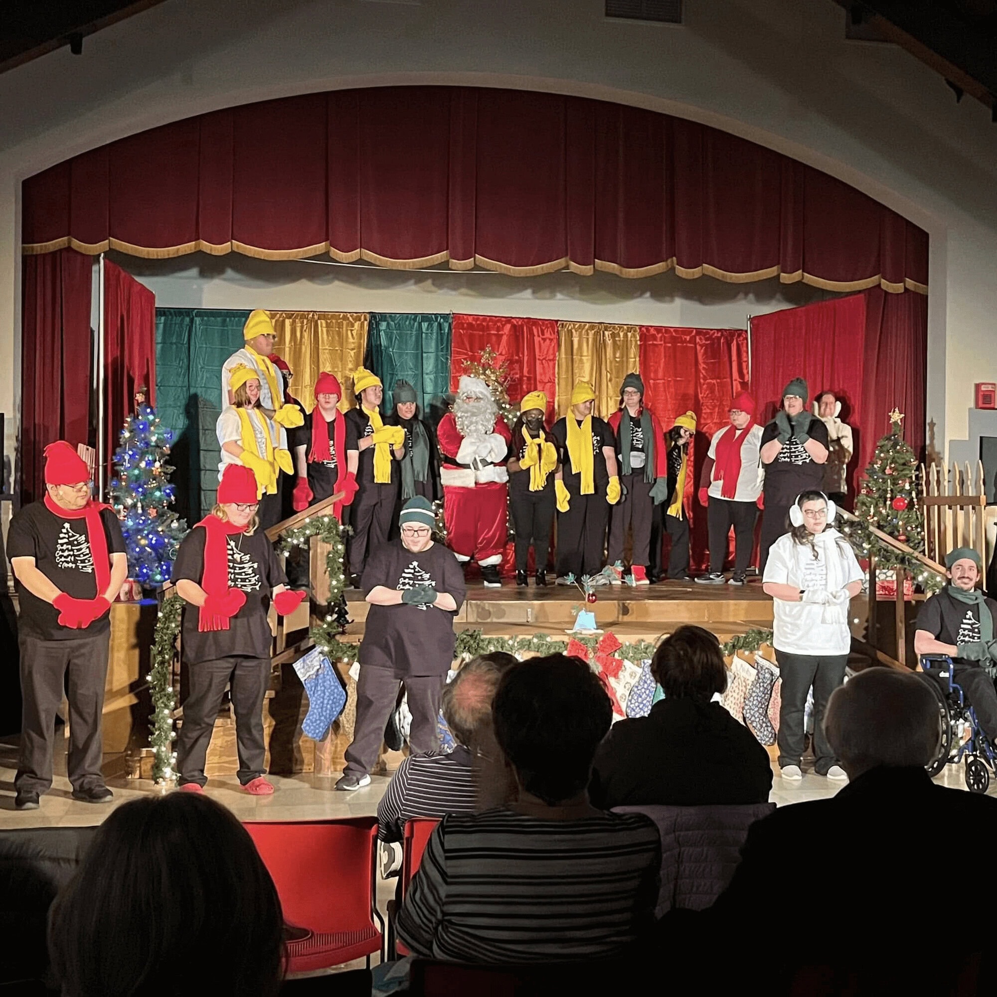 A group of performers in festive attire stand on a decorated stage with holiday trees and banners, while an audience watches from seated rows.