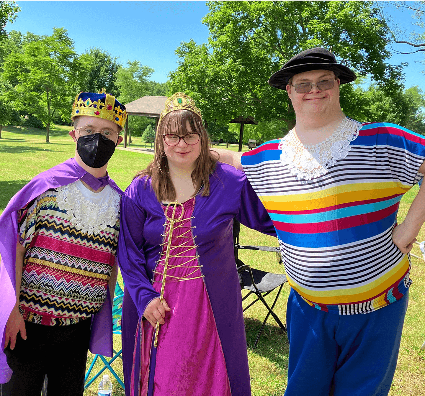 Three people in colorful costumes, including a king, a queen, and a sailor, stand together outdoors on a sunny day with trees and grass in the background.